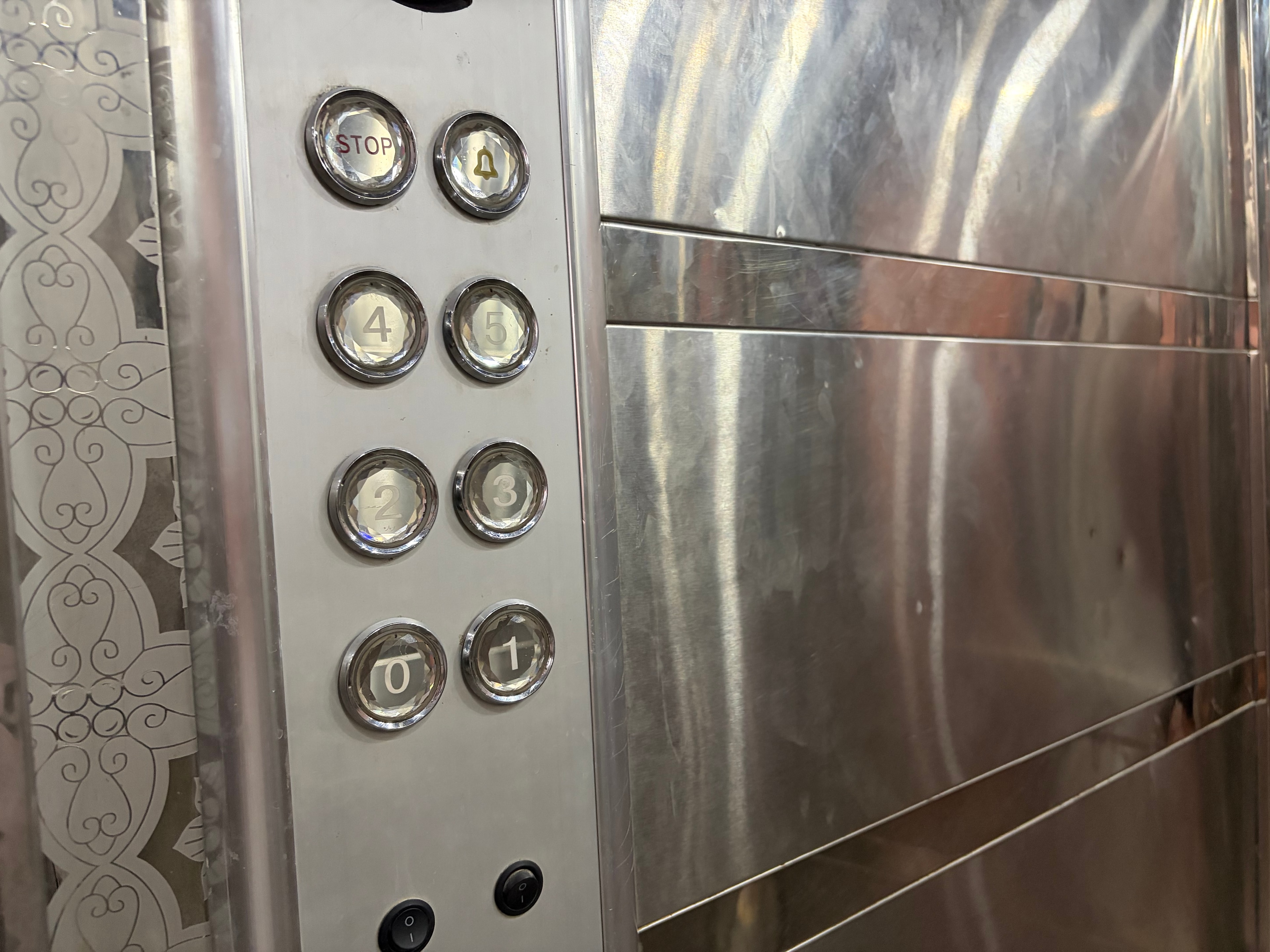 Lift control panel — floor buttons 0–5 and alarm, scratched stainless walls, etched glass mirror panels