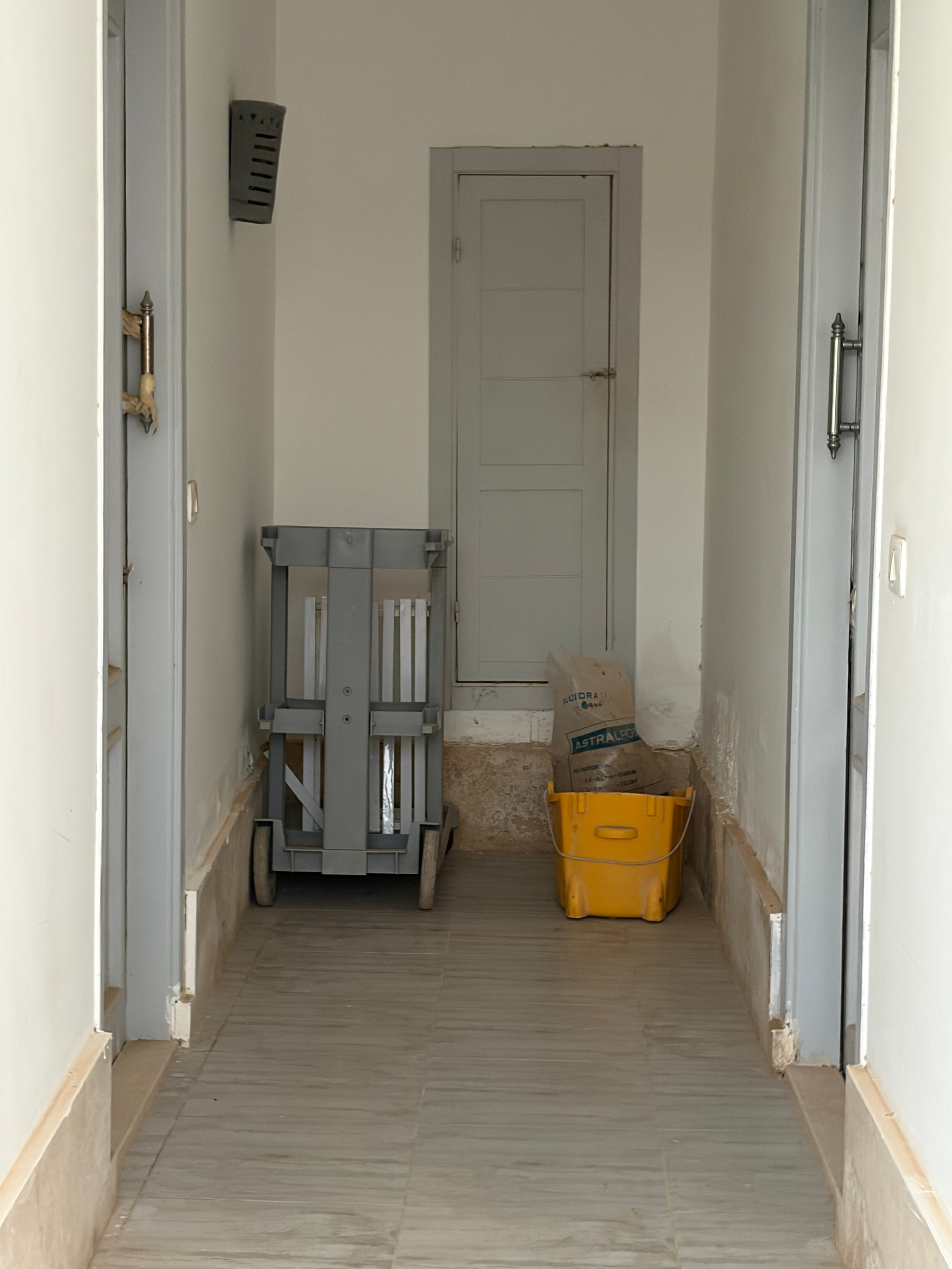 Construction materials stored in communal space — bare unfinished walls, yellow cement bucket, folding workbench and construction debris on the floor