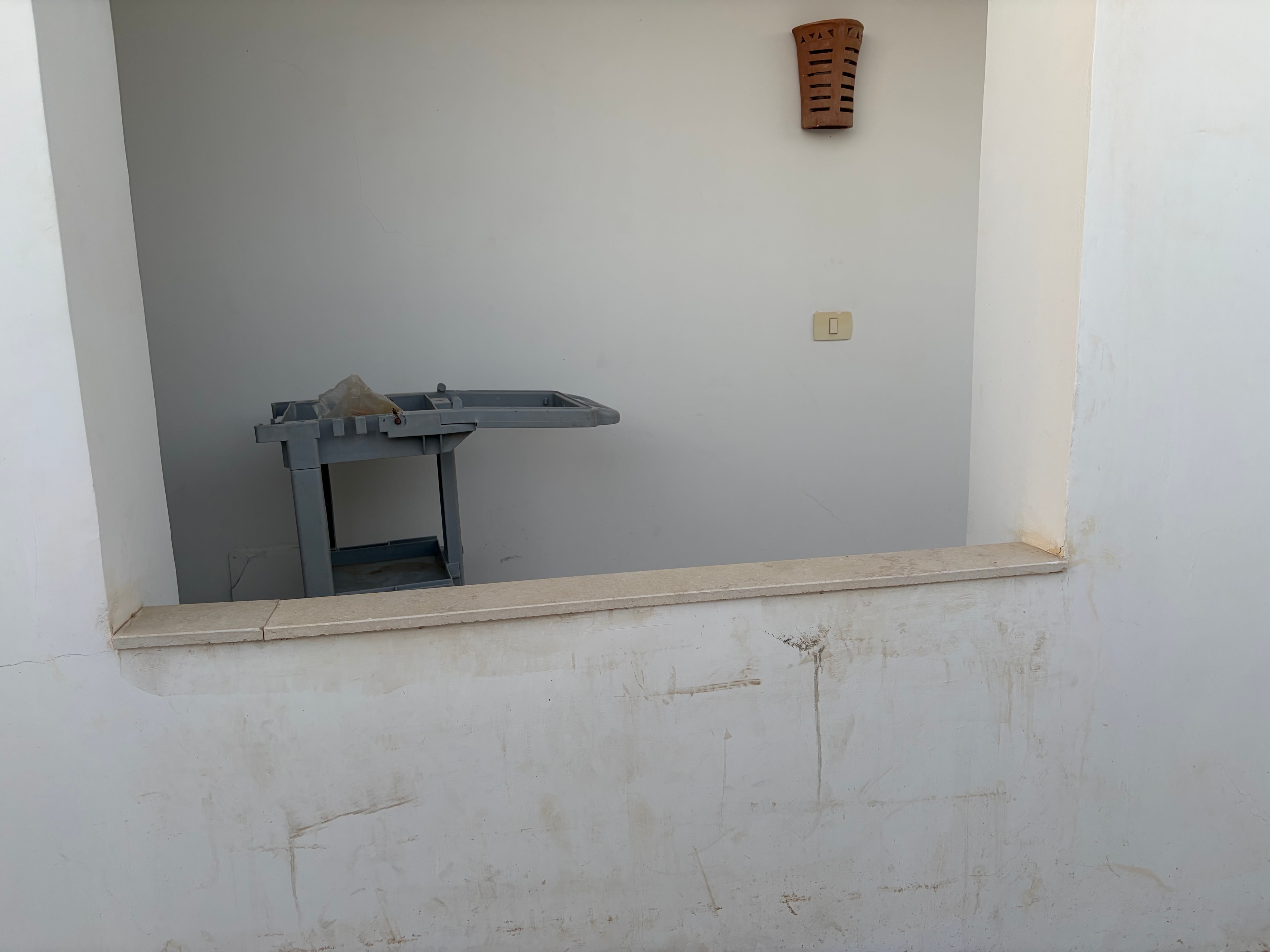 Broken AC unit hanging from ceiling — grey unit with casing open and broken, hanging at an angle above a doorway, wall above heavily stained, light switch and terracotta vent grille below