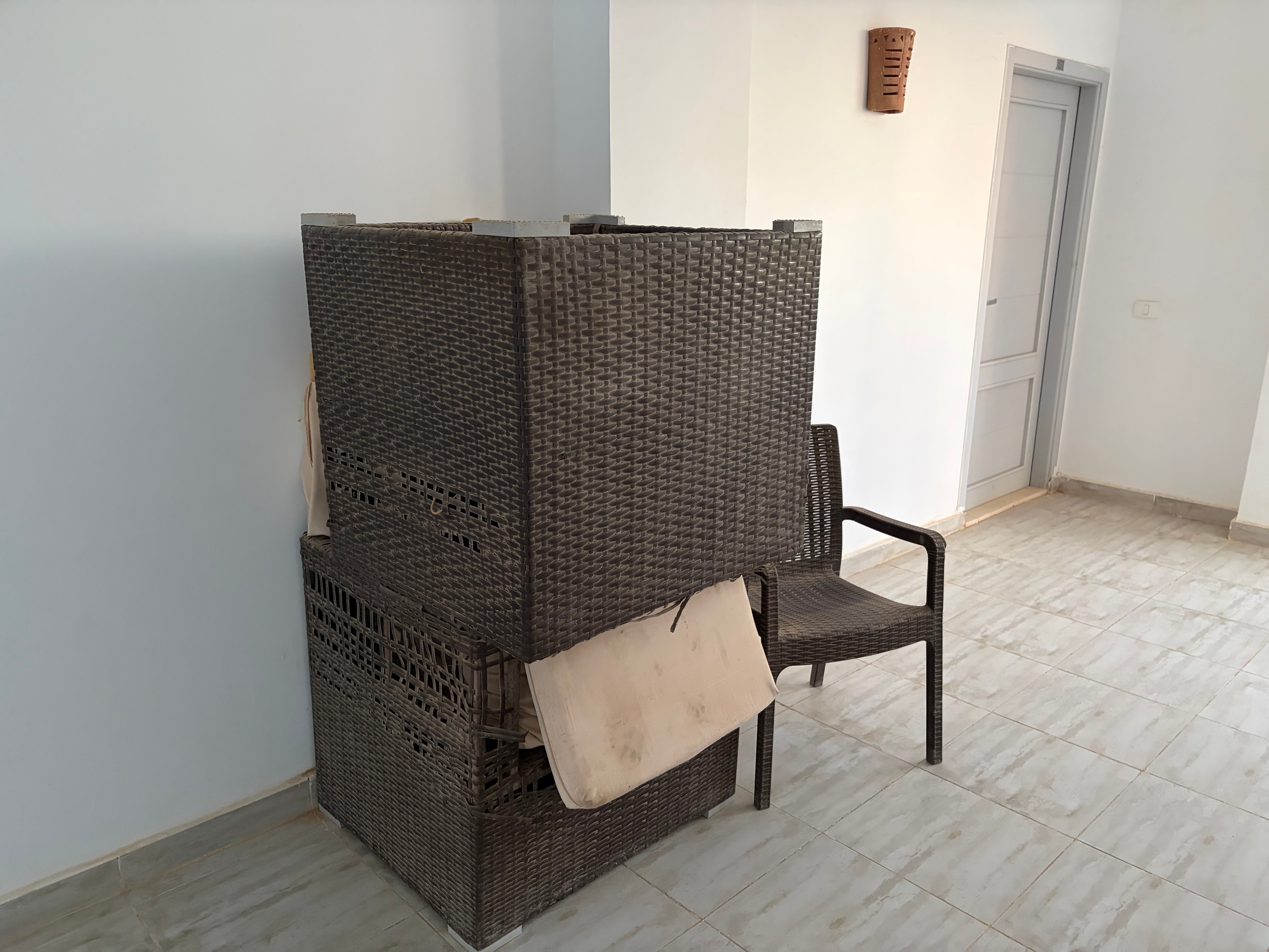 Rattan furniture stacked in communal room — multiple wicker chairs and sofa unit piled haphazardly, bare concrete ceiling with no finishing, pool area visible through window
