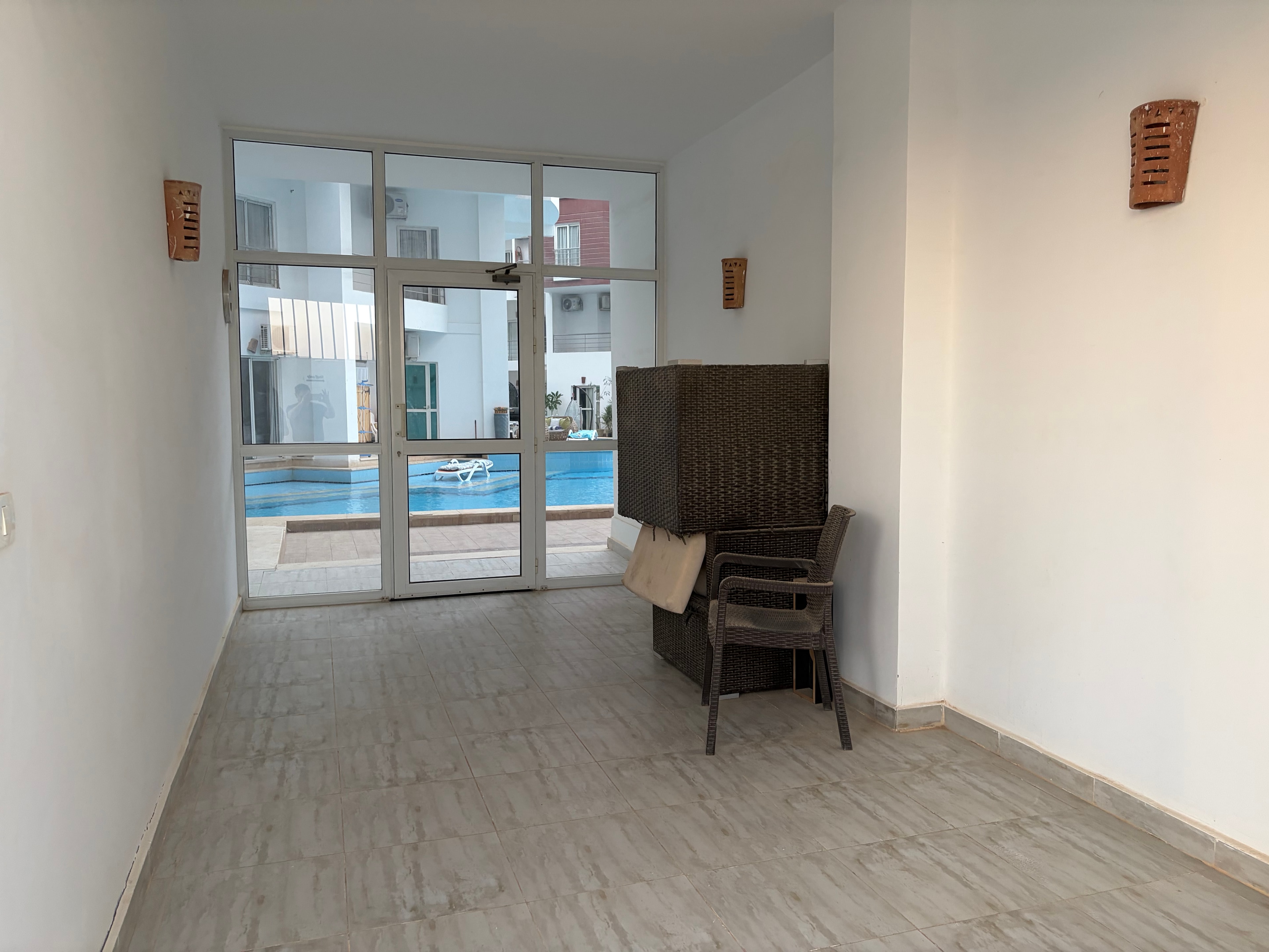 Wide shot of furniture-storage room — rattan furniture piled to the ceiling, bare concrete throughout, large windows looking out to the pool, communal space used as a dumping ground