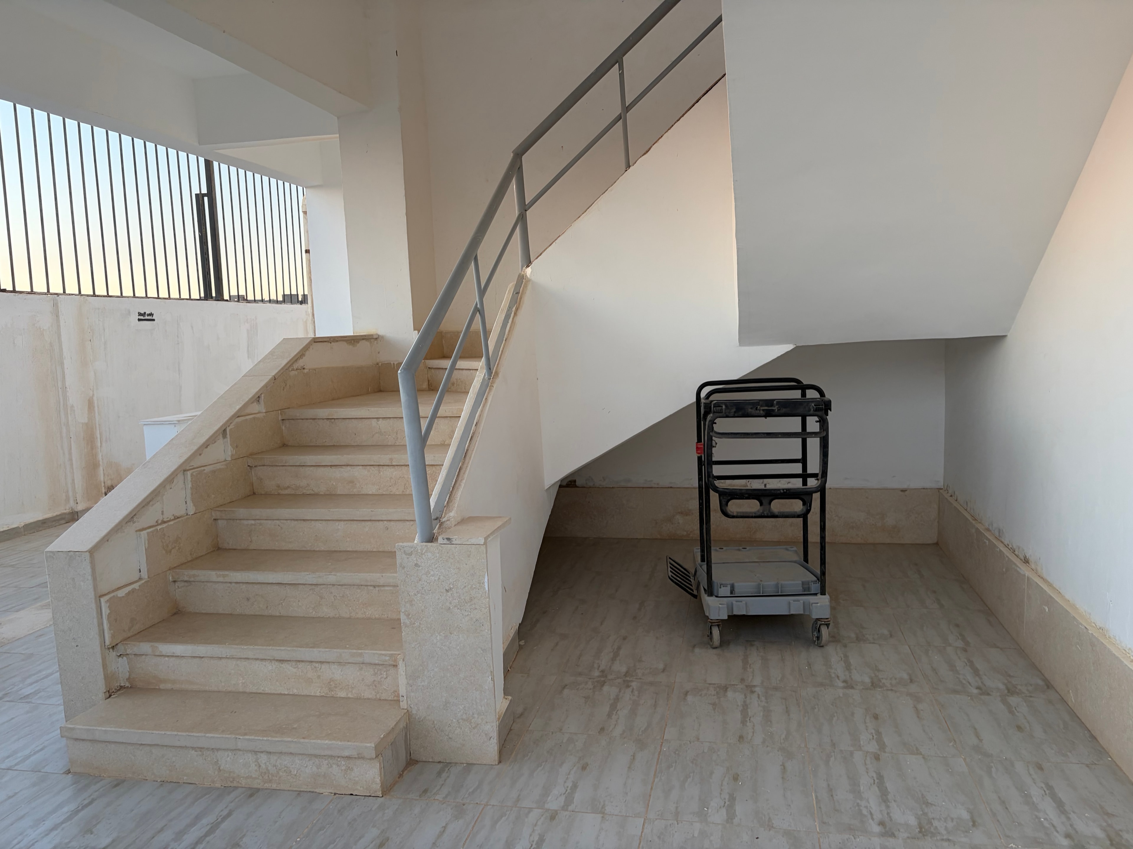 Stairwell with luggage trolley abandoned on landing — bare concrete ceiling, black metal trolley left on the landing, stairs bare unfinished stone with no cladding, simple metal tube handrail