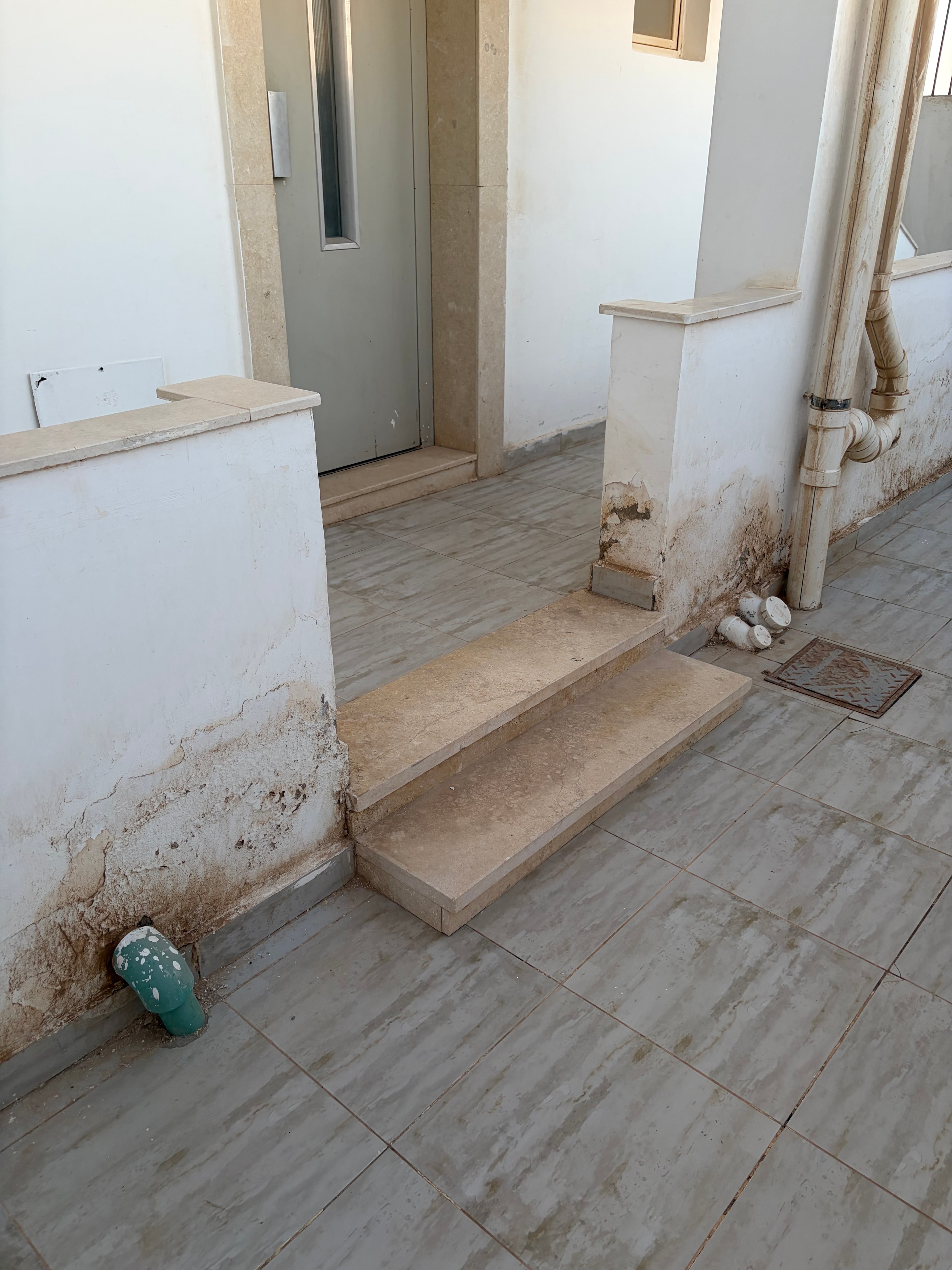 External terrace with severe water damage — extensive brown staining across tiles and down walls, crumbling render exposing substrate, exposed pipework, broken ceramic ornament, green plastic fitting on floor