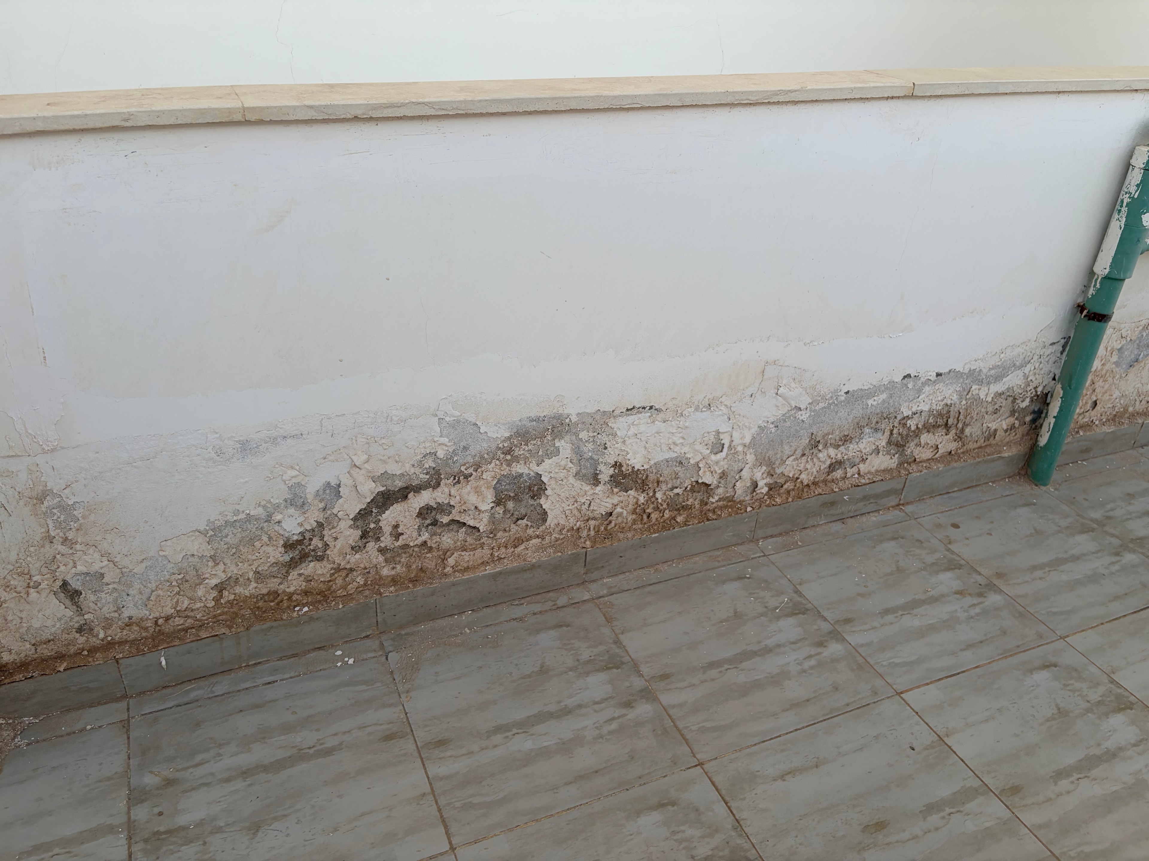 Corner junction render failure — render completely blown and crumbling at wall/ceiling corner, green pipe stub protruding from failed wall, debris on ledge below, tiled ceiling heavily stained above