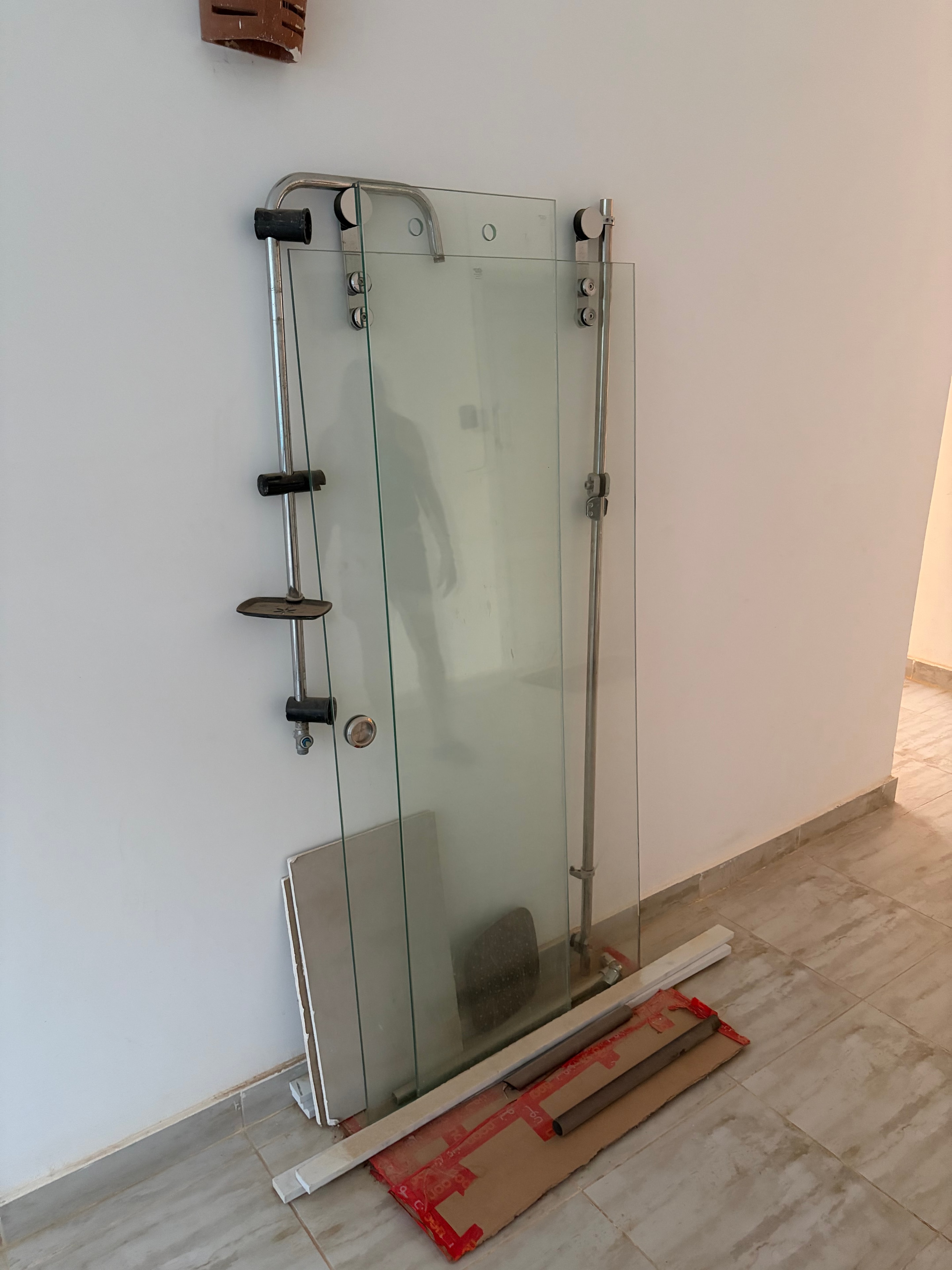 Glass shower door left on communal floor — full frameless glass shower enclosure with stainless steel fittings lying flat on the floor, cardboard packaging and building materials propped against tiled wall behind