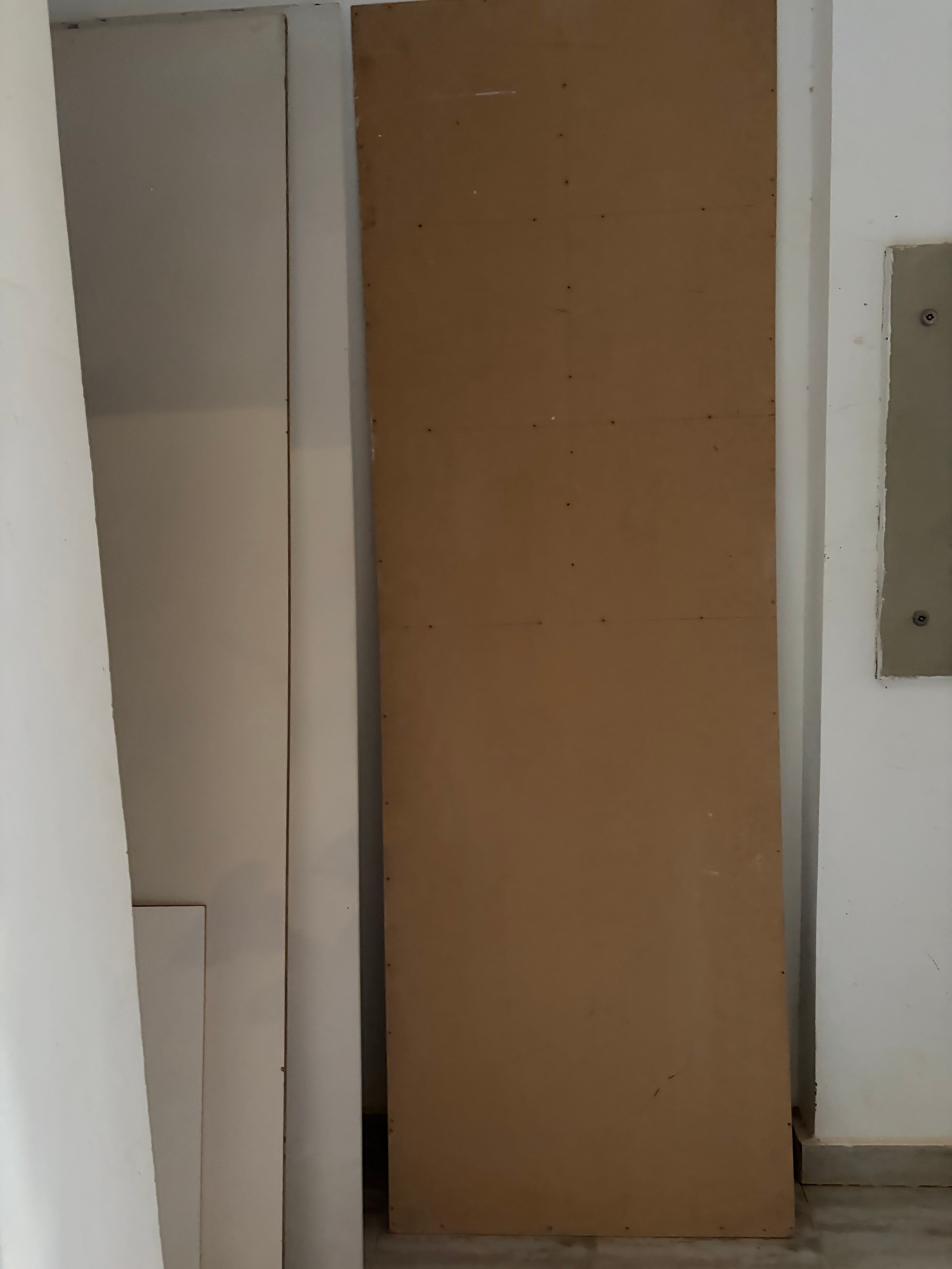 MDF panel covering wall section — large sheet of raw MDF/chipboard screwed to wall covering a window or opening, smaller grey panel below, completely temporary and unfinished fix in communal area