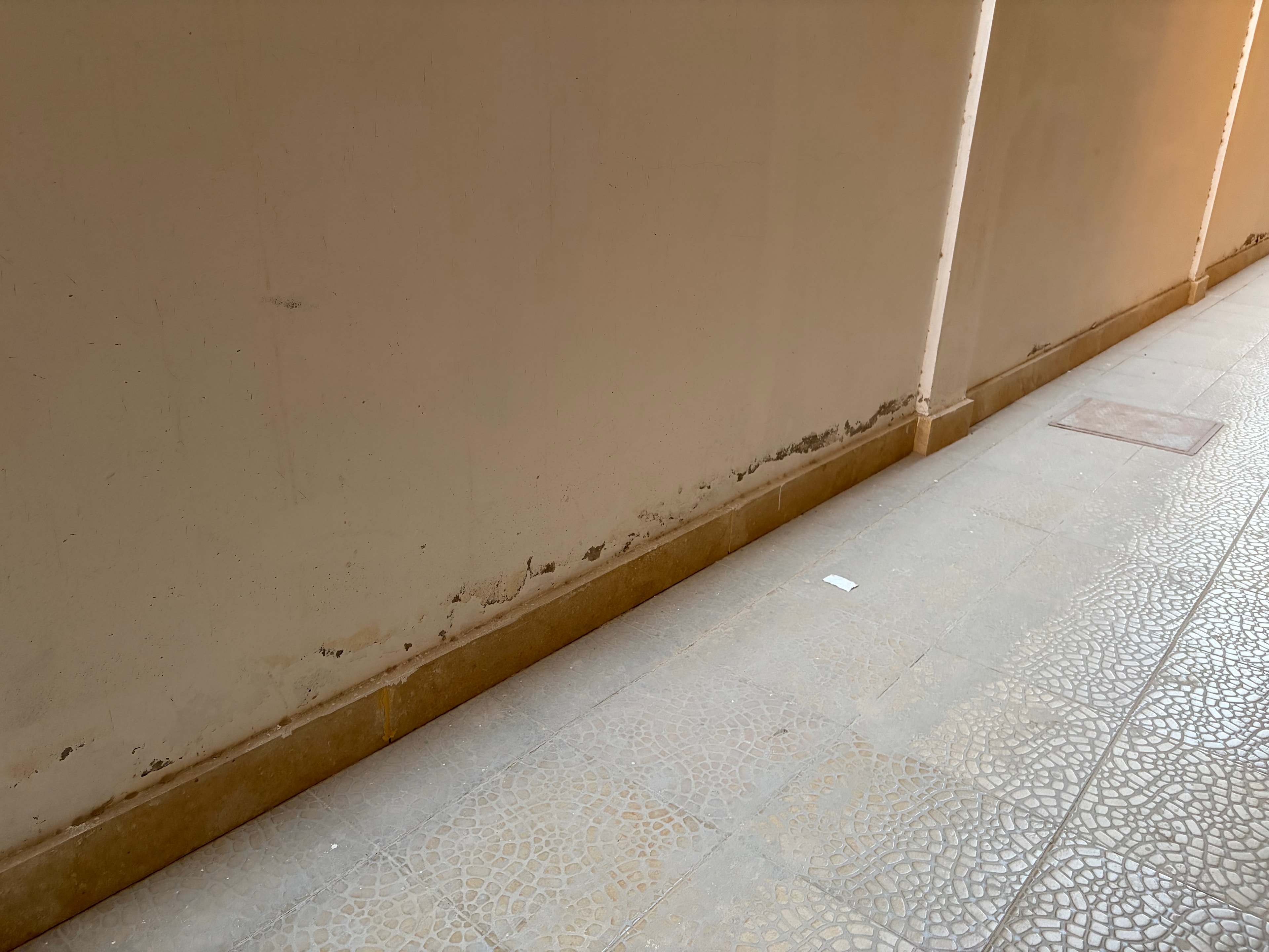 Render failure full corridor width — crumbling band running continuously along entire wall/ceiling junction, recessed lights in ceiling, vertical pipe chases on wall, scale of failure unmistakable