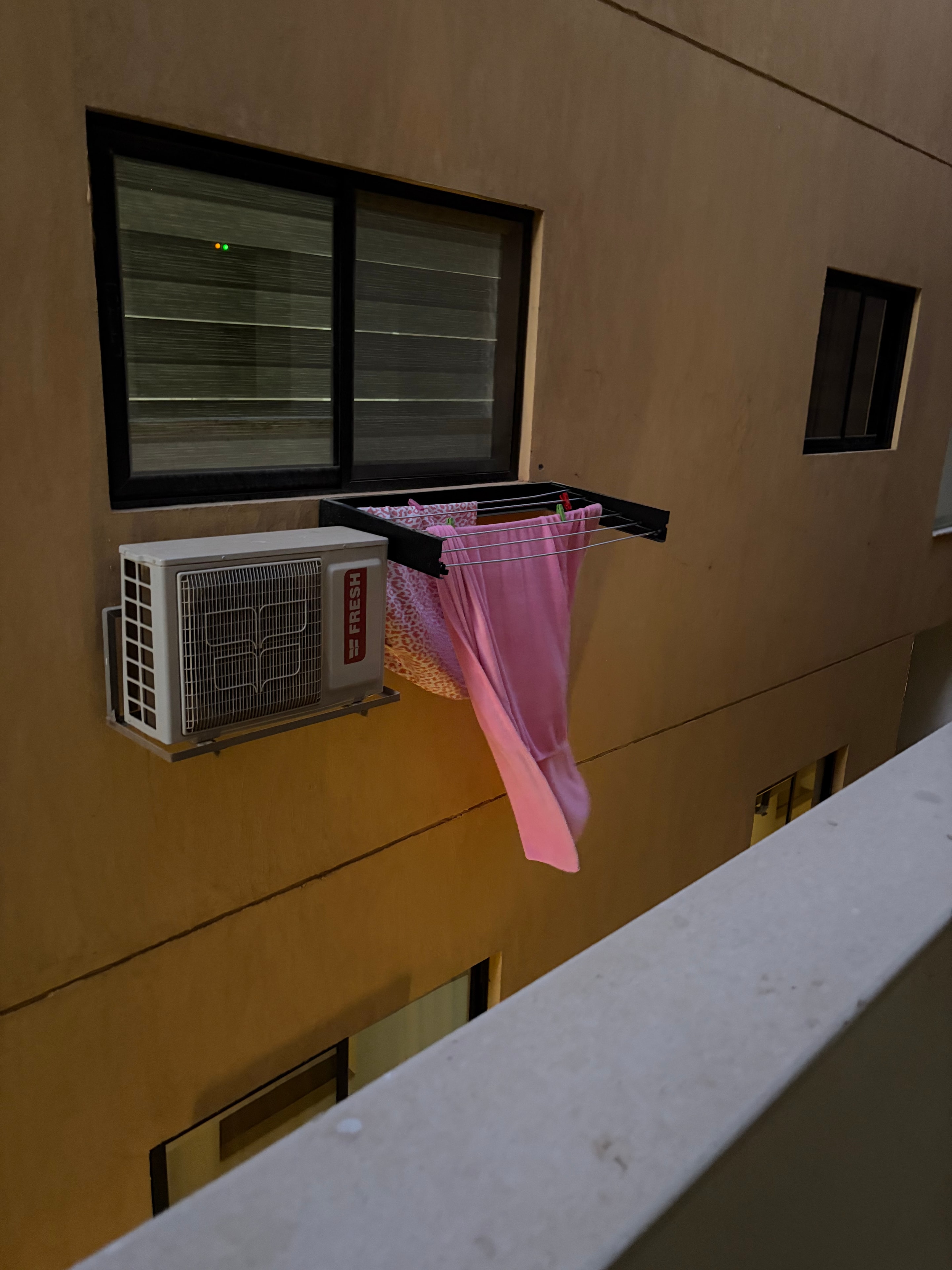 Laundry hanging from a window — pink fabric on a drying rack bolted to the building facade, FRESH brand AC unit mounted directly below, visible from communal areas