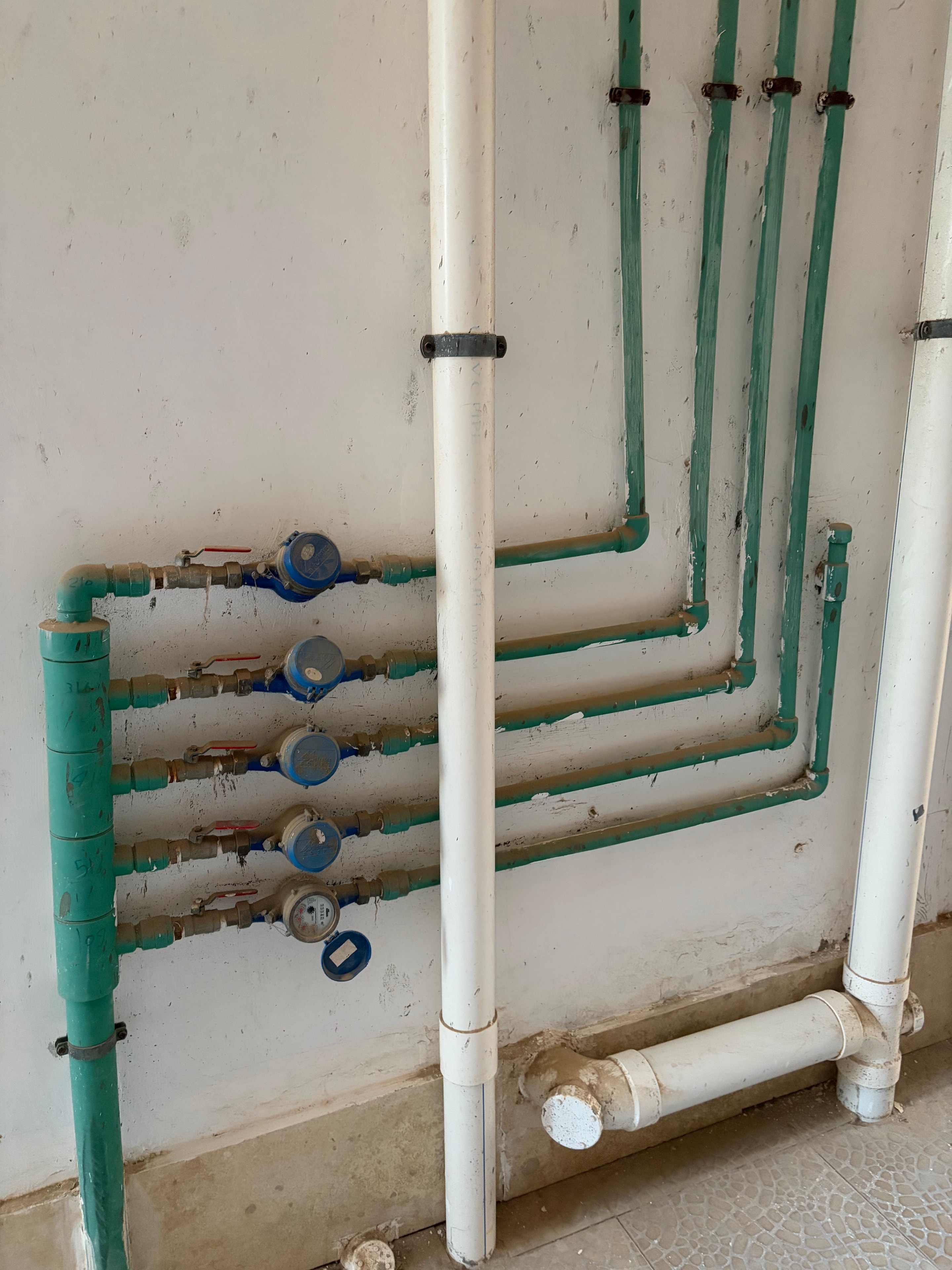 Exposed surface-mounted pipework and water meters — multiple green water supply pipes running across dirty white wall, four individual water meters with blue dials and red isolation valves all unenclosed, wall heavily marked throughout