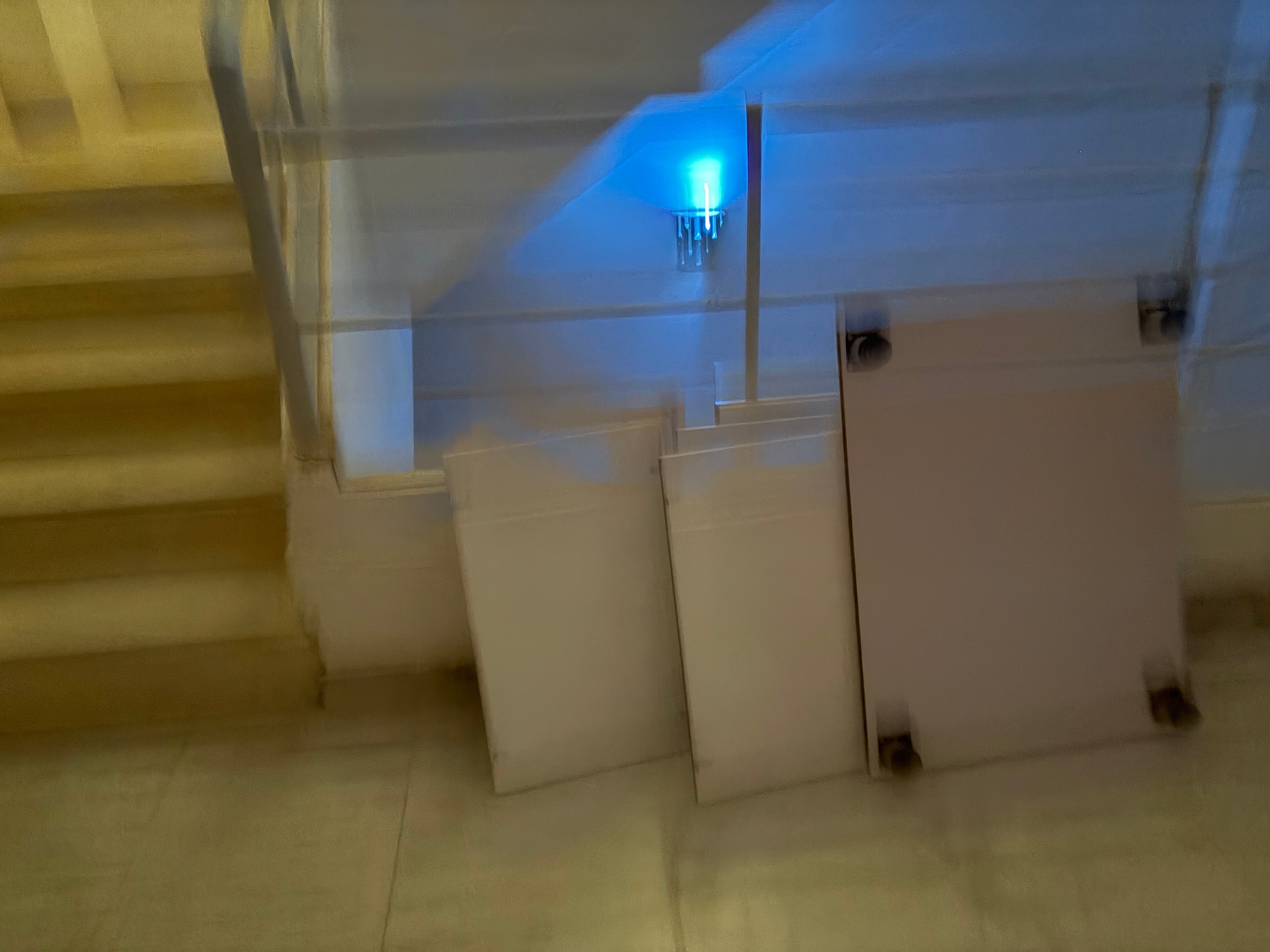 Communal stairwell lit by a single bare blue LED bulb — the only light source, staircase and walls showing heavy yellowing and staining throughout