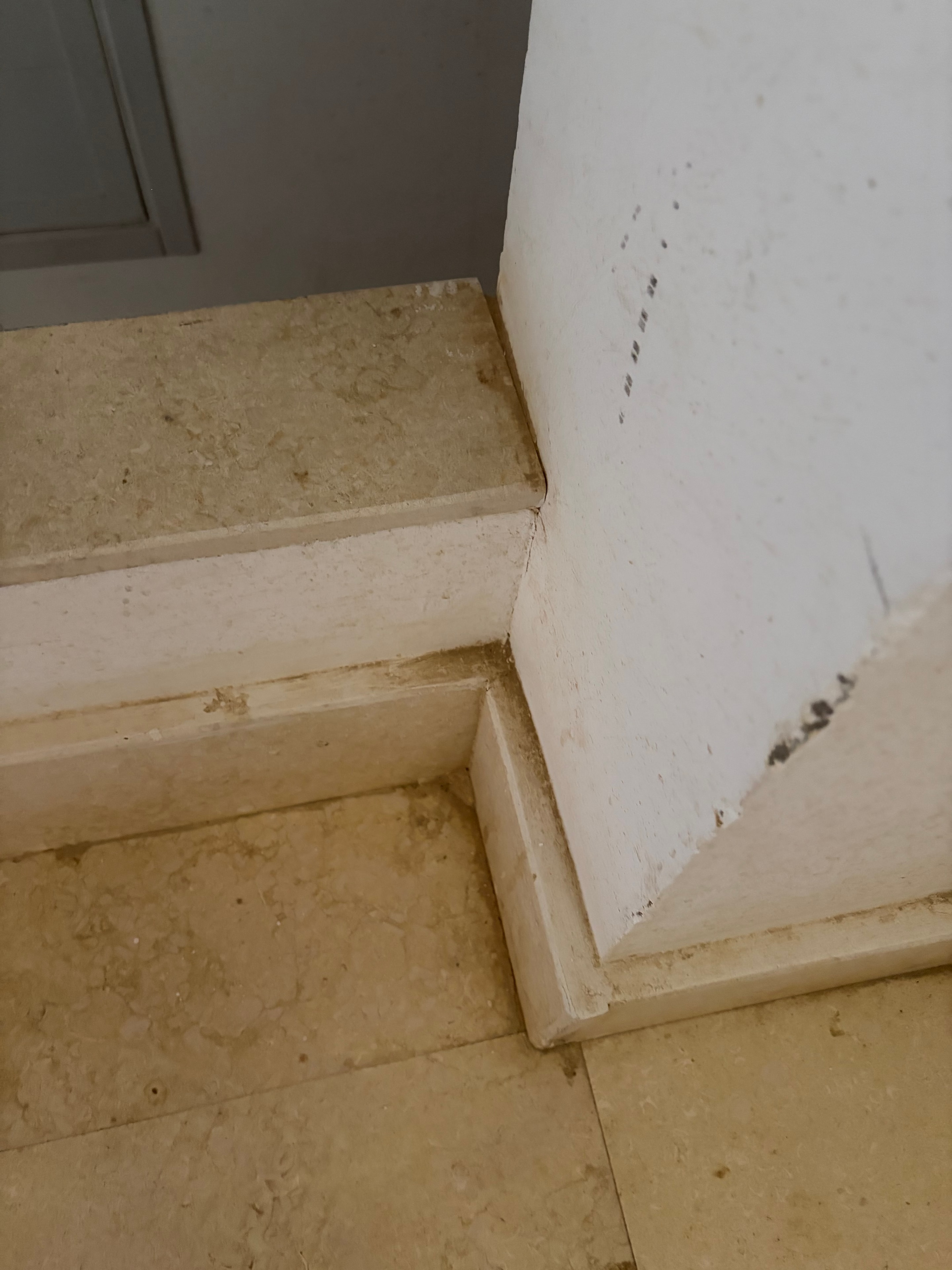 Travertine column corner close-up — heavily soiled travertine cladding at ceiling junction, black mould spots on white ceiling above, cornice detail heavily marked and dirty, filth accumulated in every joint and recess