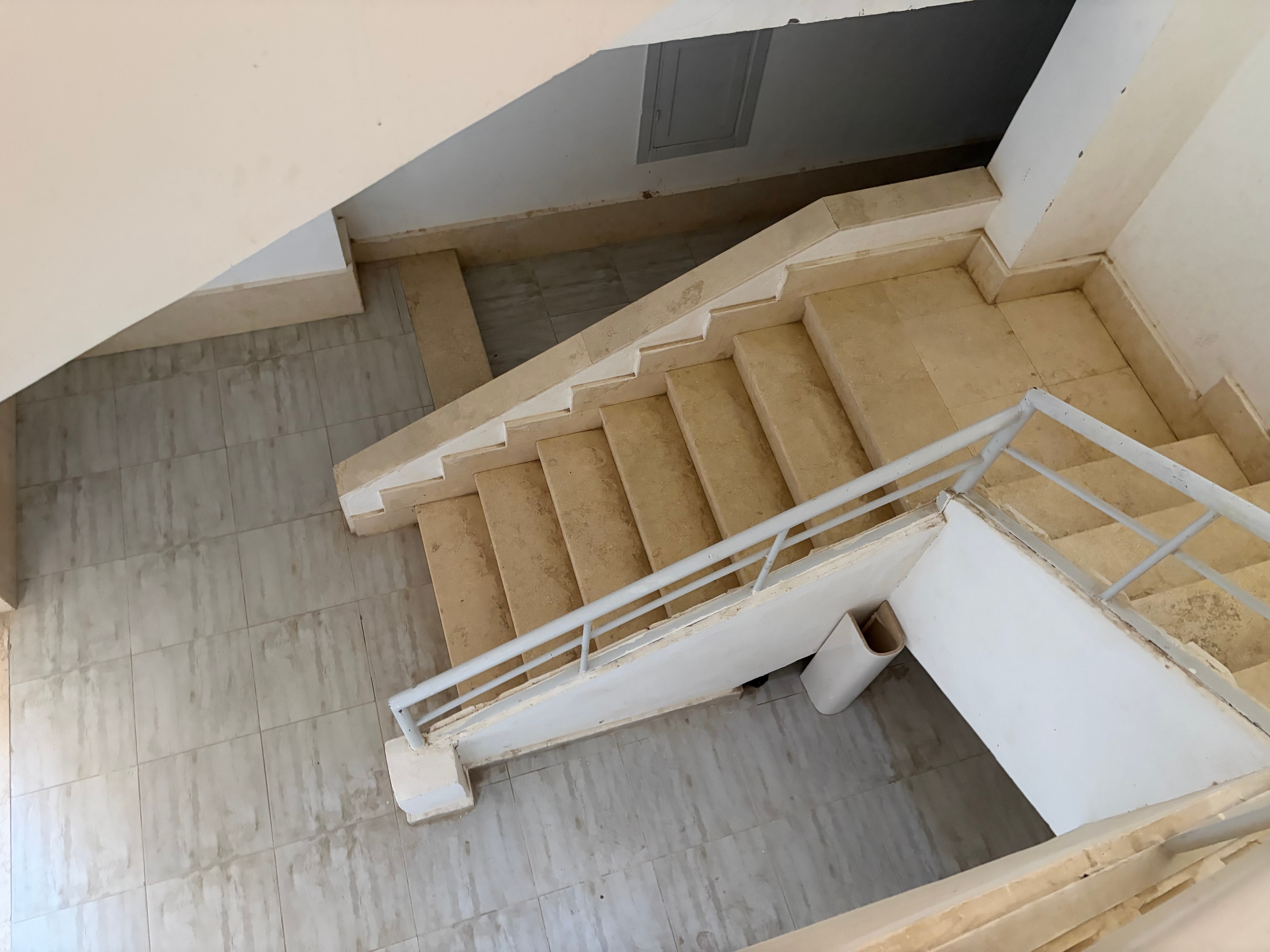 Stairwell looking straight up — multi-storey travertine staircase, large white PVC pipe abandoned on a landing above, stairwell walls heavily stained throughout, grey fire door at bottom