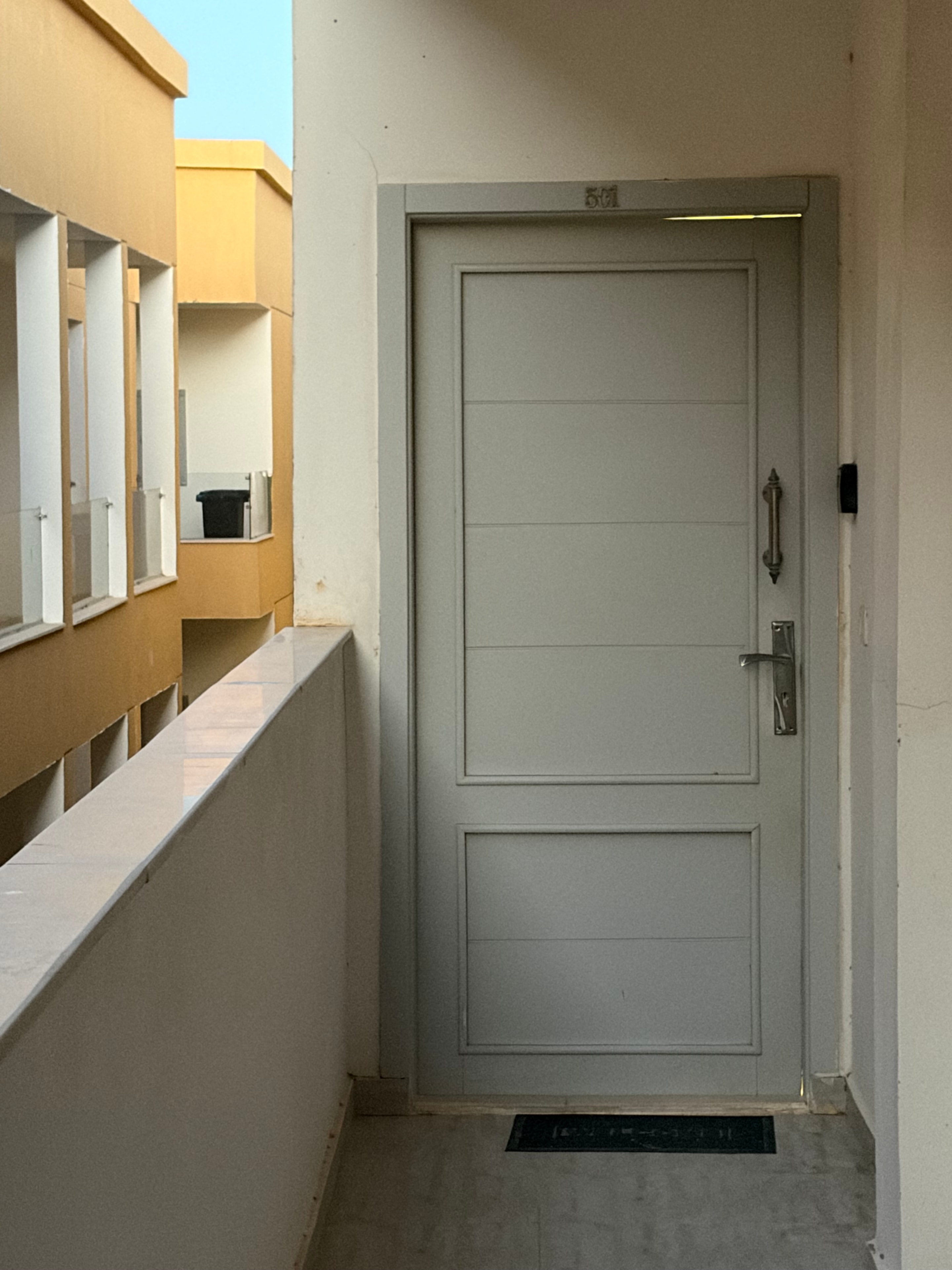 Unfinished communal space — large white panelled shutter set into a wall above a ledge, open atrium below showing unfinished yellow render facade and multiple AC units